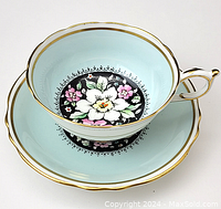 Side angled view of Paragon cup resting on matching saucer showing floral design with white, pink flowers on black background inside light blue cup and saucer with gold trim.