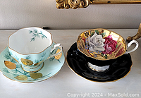 Two porcelain tea cups and saucers with Queens branding, featuring gold leaf and floral designs, side by side on a white surface.