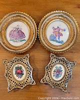 Four framed needlepoint artworks placed on wood surface: two oval gold-tone dancing figures and two silver-tone floral lattice frames.