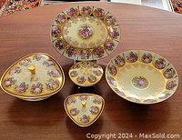 Top view of the full collection showing oval plate, round bowl, and three covered bowls of triangular and round shapes featuring painted romantic scenes and gold trim.