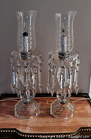 Pair of crystal luster lamps with their glass hurricane shades and hanging crystal prisms on a polished wooden surface, unplugged.