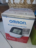 Photo of the Omron 6 Series Upper Arm Blood Pressure Monitor box showing branding, features, and a display image of the device with blood pressure readings on an LCD screen.
