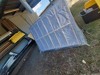 Box spring wrapped in plastic covering, leaning upright outdoors against a metal-roofed structure.
