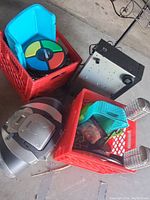 Photo shows two red plastic milk crate bins containing various items including a Simon electronic game, a portable boombox stereo, a small lockable safe, a turquoise plastic chair, coiled cords, and other assorted household and decorative items. The photo captures an 80s and 90s theme bin mix.