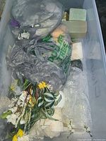 Clear plastic bin showing assorted artificial flower stems, floral foam blocks, plastic bags, and bubble wrap inside.