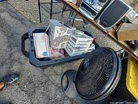 Photo showing a large black oscillating floor fan in front of a plastic storage bin containing various boxed and loose decor items including small fans or lighting fixtures and white slatted items.