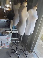 Photo shows two white female mannequins on black metal stands with caster wheels positioned in a storage or garage setting, partly illuminated by daylight coming in from a door.