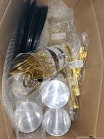 Black plastic plates stacked inside a cardboard box with some forks and clear plastic cups packed together.