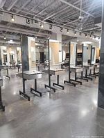 Wide view of multiple double sided salon stations in a commercial space. The lot is one of these stations, featuring large mirrors with lights, black metal bases, and countertops with drawers.
