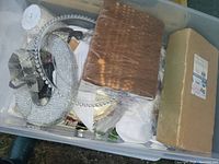 Bin containing various wedding decoration items including silver rhinestone circle, brown napkins or fabric pack, and other decorative pieces.