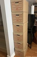 Stack of six cardboard banker file storage boxes with attached lids and printed labeling on front sides.