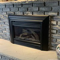 Front view of gas insert fireplace unit installed in brick hearth with black metal surround and ceramic logs visible through glass.