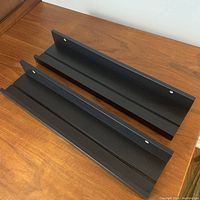 Two black IKEA Mosslanda picture ledges shown on wood surface, displaying front and angled top views.