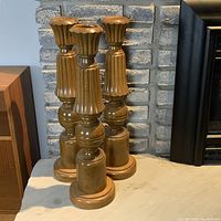 Front view of three tall wooden candle sticks placed on a light-colored surface in front of a gray brick wall.