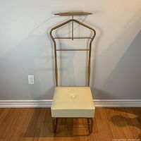 Full view of the mid-century valet stand with padded cream seat, gold-toned metal frame, wooden tray, and shoe rack, shown against wall and hardwood floor.