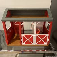 Red plastic play barn with a gray roof. Visible closed half doors forming two stalls and open tack room inside with hooks and a hanging orange bucket.