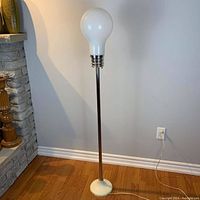 Floor lamp standing on hardwood floor next to a stone fireplace, showing the full height and shape with the large white frosted glass bulb on top.