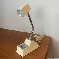 Front view showing the lamp upright on a wooden surface with cream-colored base, rectangular light head, adjustable metal arm, and the switch panel with two settings.