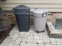 Both garbage bins side by side on pavement outside a brick building; black bin on left, beige bin on right with lid and wheels.