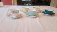 Five Aynsley cups and saucers displayed on white cloth showing variety in color and design