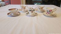 Five porcelain cups and matching saucers displayed on a white tablecloth, showing variations in patterns and colors.