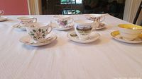 Wide angle photo showing all six cups and saucers arranged in a row on a white tablecloth with various floral and scenic designs.