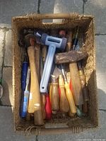 Basket filled with various hand tools showing handles and tool heads including hammers and files.