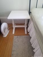 Front side view of the white bedside table placed next to a bed and small fan on wooden floor.