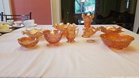 Full view of all 9 carnival glass pieces arranged on a white tablecloth, showing variety of shapes and sizes with ruffled edges and iridescent orange finish.