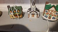 Two ceramic Christmas village light-up buildings on a gray surface: a yellow house with green roof and a white church with clock tower, each with attached power cords.