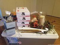 Photo showing 4 boxes of 100 white string lights stacked, artificial flower arrangement with pine cones and berries, elf decoration, and clear plastic or glass items on top of a large white storage container.