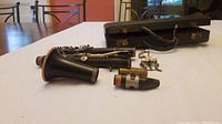 Photo showing disassembled black clarinet body parts with silver keys and a mouthpiece with ligature, all placed on a table with case in the back.