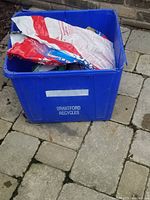 Blue recycling box filled with packets and bags of de-icer and salt shown from above on paving stones.
