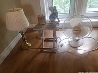 Full view of five items: folding table, brass lamp, brass clock, porcelain figurine, and white fan on wooden floor near window.