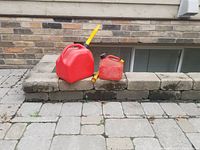 Two red gasoline gas cans sitting side by side on stone steps outdoors; large can on left, smaller can on right with yellow sprout attached to larger can