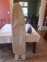 Photo showing the ironing board with a floral cover in a room setting, standing upright with table and chairs in background.