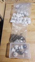 White ceramic knobs in sealed plastic bag, dark metal handles in adjacent plastic bag, and a plastic container containing more knobs and handles on a wooden surface.