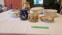 Wide photo of lot items on table: Fitz and Floyd floral pitcher, Denby casserole, souffle dish, blue cow jar, ceramic floral piece, cheese plates.