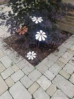 Four metal rustique flowers planted in garden soil surrounded by light-colored paving stones. Flowers have rust-colored petals with a round rusted center. Three large flowers and one small flower clustered together.