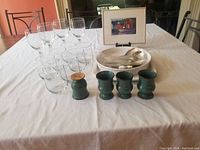 Table with grouped clear wine glasses, tumblers, green ceramic cups, large metal bowl, and framed art print on black display stand.