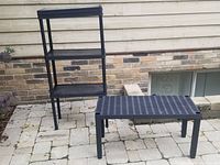 Photo showing the three-tier black plastic shelving unit next to the low rectangular stand outdoors on stone paving against a brick background.