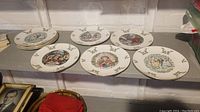 Nine Christmas plates displayed on a surface, showing various illustrated holiday scenes and holly decorative edging.