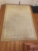 Photo of neutral beige rectangular area rug with a simple double border on hardwood floor. Rug shows dirt, stains, and discoloration, needing cleaning.