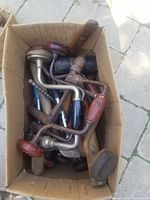 Inside view of a box filled with assorted vintage hand braces and small hand tools. Mostly metal braces with red, black, and wooden handles.