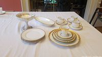 Wide shot of the white and gold china set arranged on a white tablecloth showing cups, saucers, plates, side dishes, and serving dish.