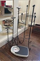 Four metal candle holders of varying heights with decorative bases next to Metro brand mechanical scale with white dial and black platform.