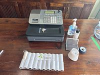Overview of Casio PCR-T285 cash register with accessories including thermal paper rolls, keys, sanitizer, lotion, tape, and small box arranged on wooden table.