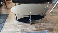 Front angle view of the oval glass coffee table showing the clear glass top, metal legs, and black glass shelf under the tabletop.