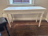 Full view of the white painted pine wood console table with turned legs and rectangular top against a wall with window above.