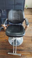 Front view of black vinyl upholstered hairdressing chair showing padded seat, backrest and armrests with chrome metal base.